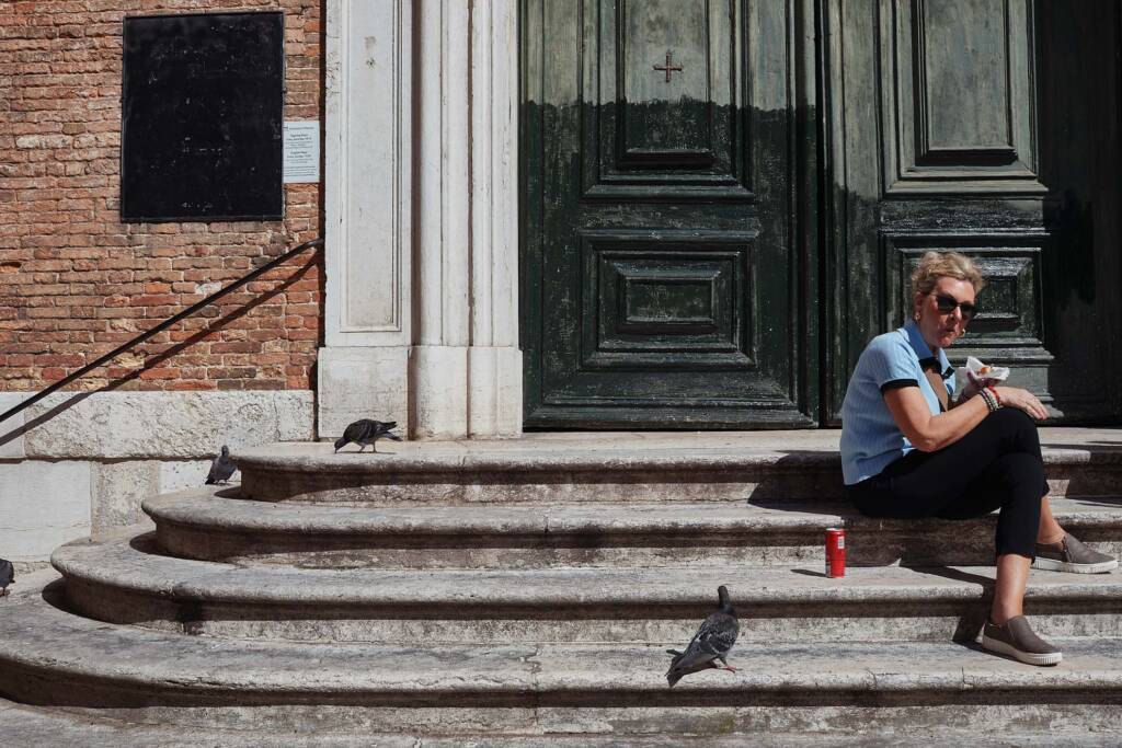 Persona seduta sui gradini di pietra davanti al portone scuro di una chiesa veneziana