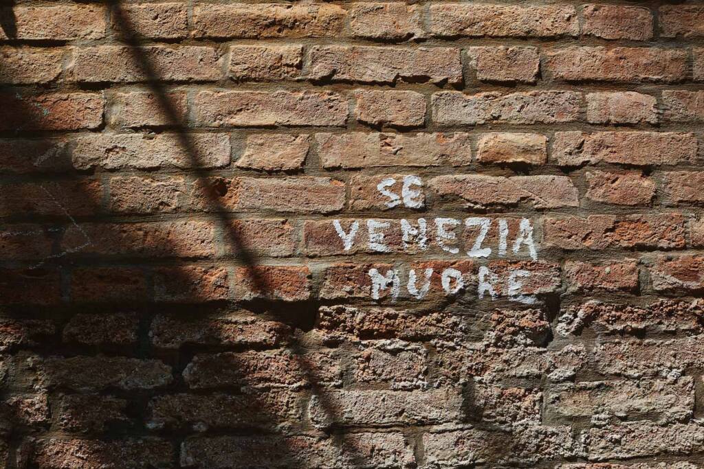 Scritta a vernice «Se Venezia muore» su un muro di mattoni illuminato dal sole