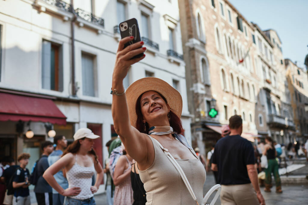 Donna con cappello di paglia si fotografa con lo smartphone in una calle veneziana affollata